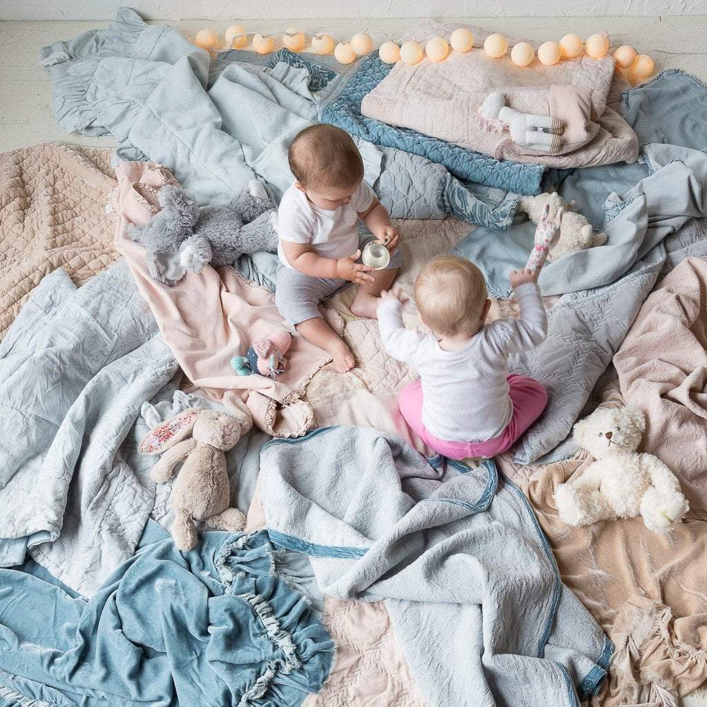 : Two babies sitting and playing on a floor covered with rumpled blankets in a variety of textures of blue, grey, and pink tones - overhead view.
