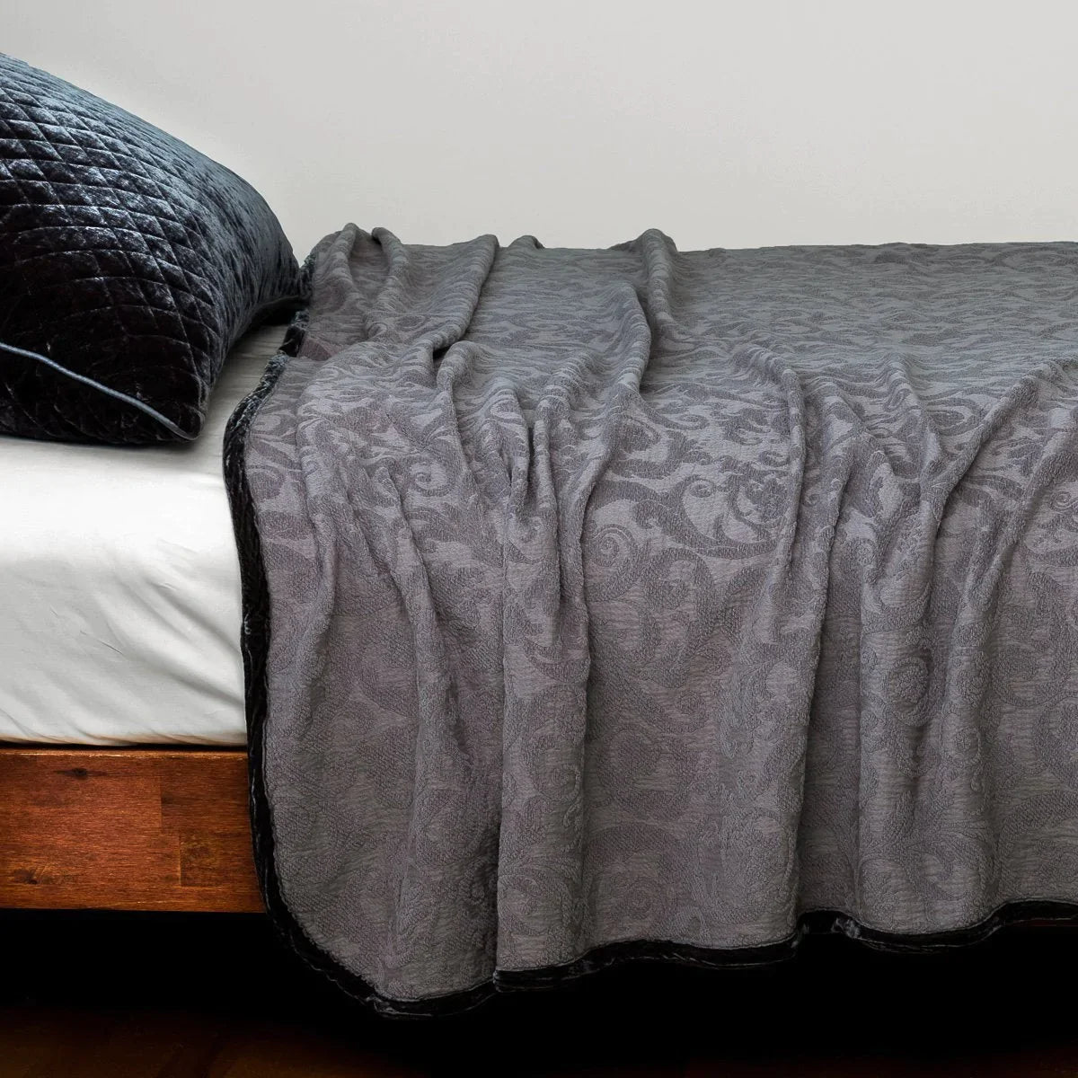 [allvariants]: The Adele coverlet in moonlight, styled on a wood framed bed shown from the side. Shown with a white sheet and a silk velvet quilted sham in moonlight.