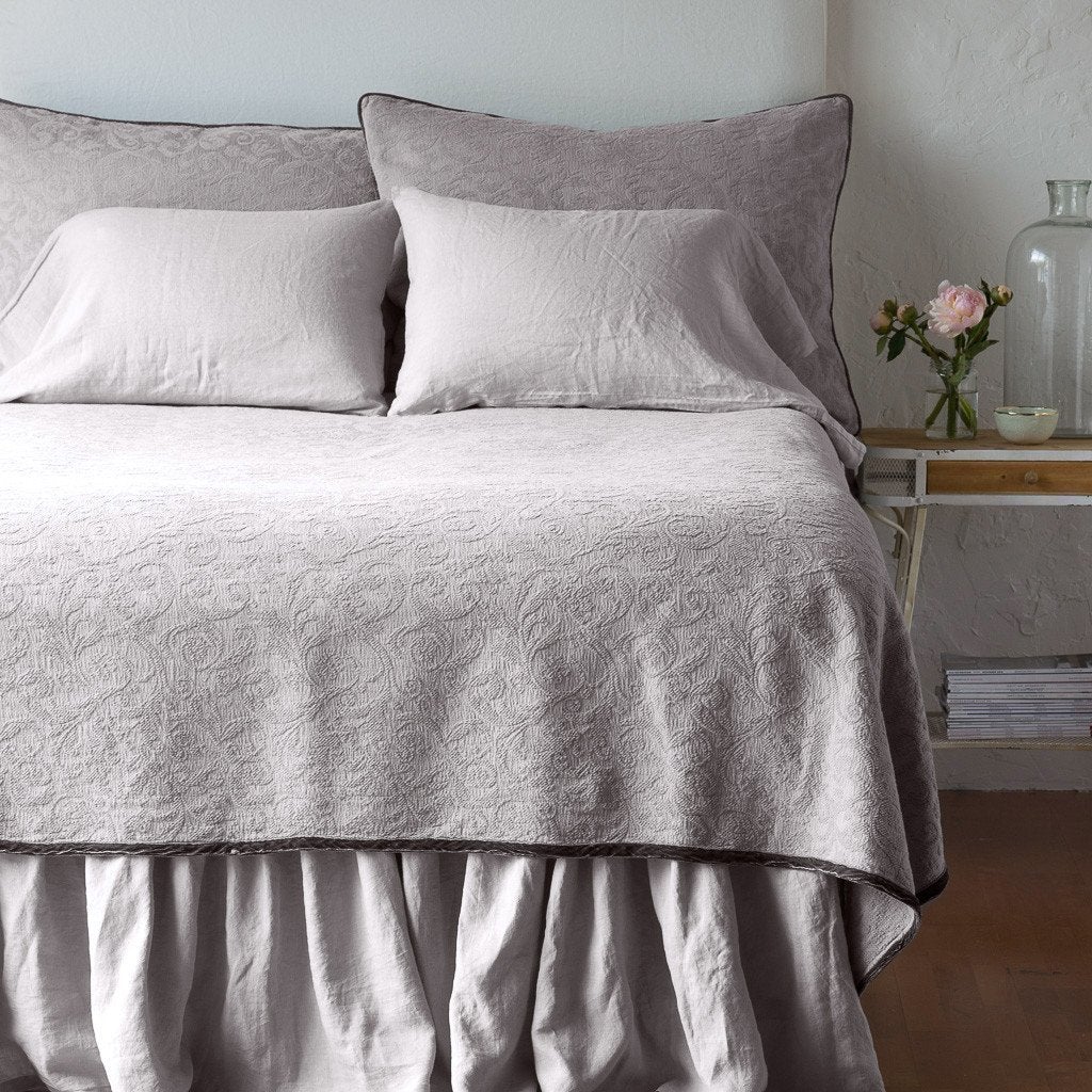 Fog: The Adele coverlet in fog, viewed from the foot of the bed, with white bed skirt and pillows visible. The headboard is plain light color, and part of a bedside table is visible with flowers, a vase and some books. against a plain white wall.