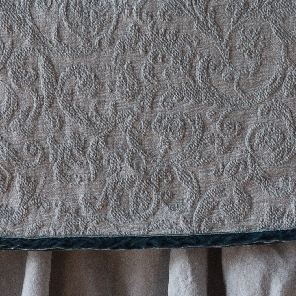 Mineral: Close up of the Adele coverlet in mineral, highlighting the detail of the cotton damask and the silk velvet edging trim. A white bed skirt peeks out at the bottom of the shot.