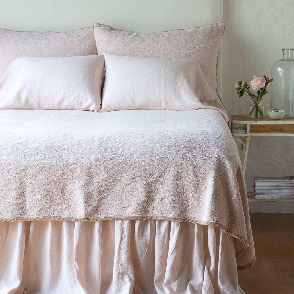 Pearl: The Adele coverlet in pearl, viewed from the foot of the bed, with white bed skirt and pillows visible. The headboard is plain light color, and part of a bedside table is visible with flowers, a vase and some books. against a plain white wall.