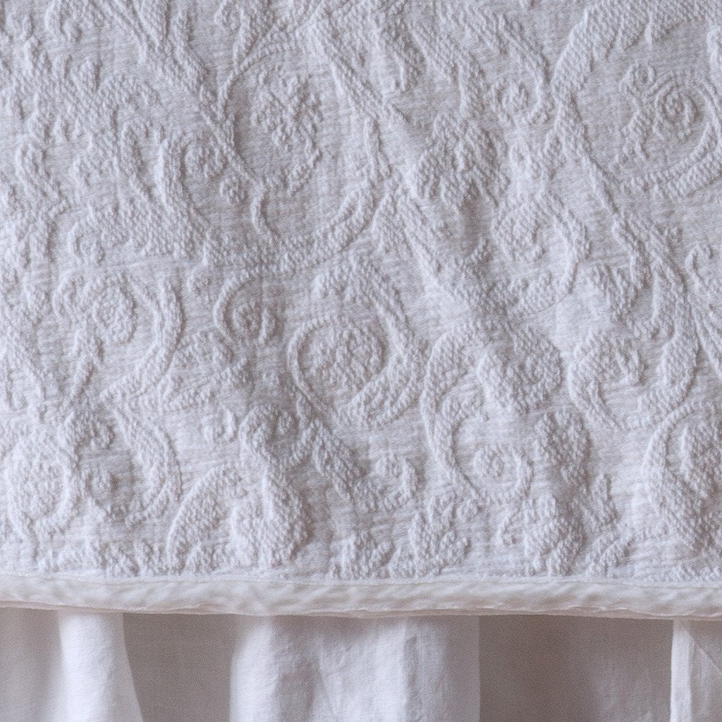 White: Close up of the Adele coverlet in white, highlighting the detail of the cotton damask and the silk velvet edging trim. A white bed skirt peeks out at the bottom of the shot.