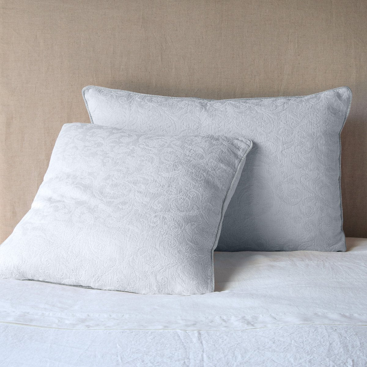 Sterling: Two Adele shams, shown in euro and deluxe sizes, leaning against a neutral toned headboard on white bedding.