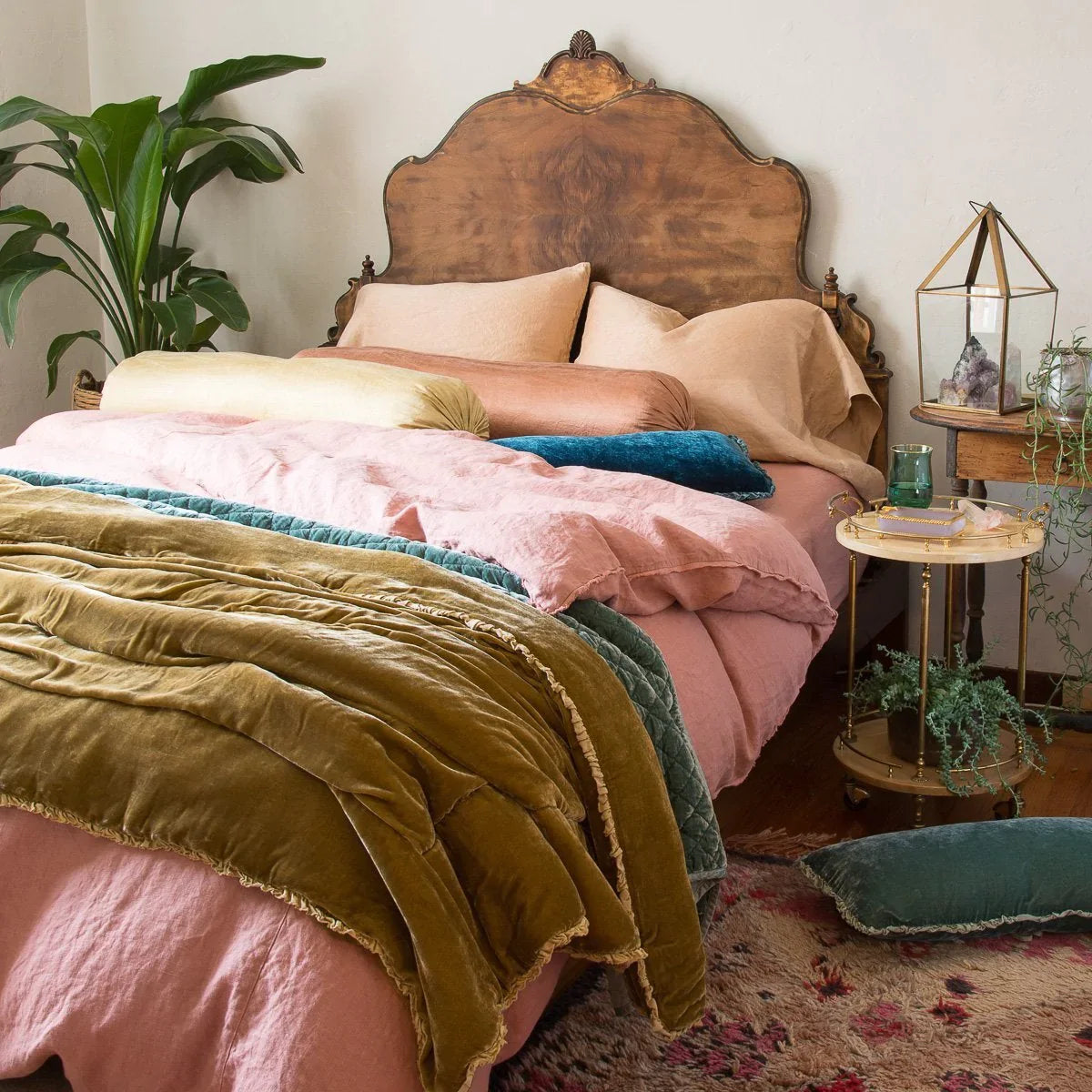 [allvariants]: Austin duvet cover shown from three-quarter angle, layered with silk velvet and silk charmeuse accessory pieces in pink, gold and blue-green tones. Dramatic wood headboard, greenery, patterned rug, and eclectic bedside styling create a bohemian vibe.