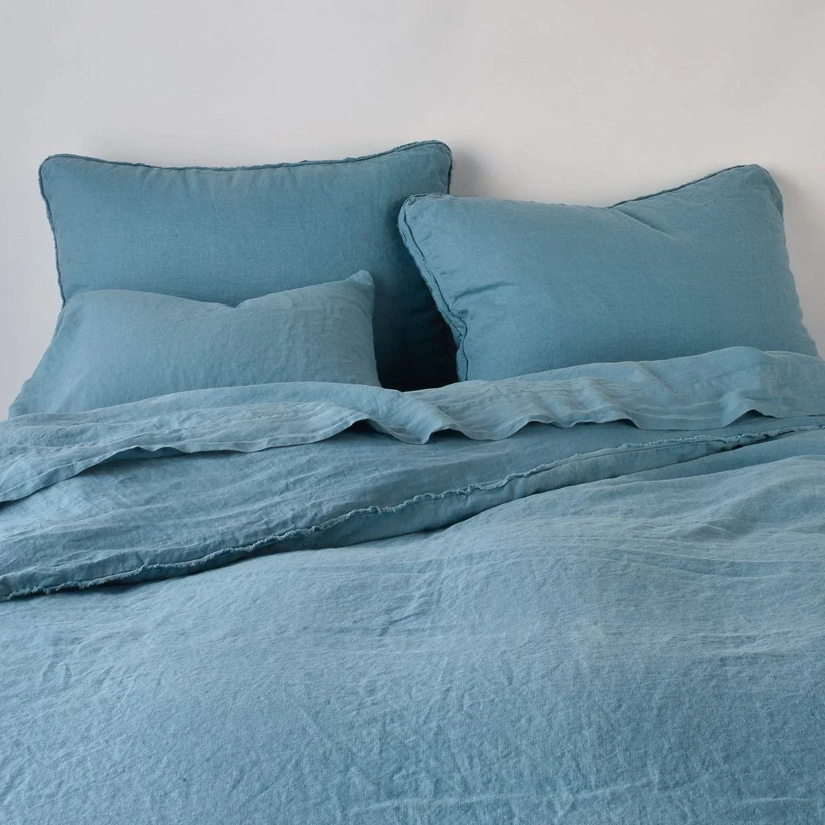 Cenote: Midweight linen shams shown from foot of bed, leaning against a plain background with monochromatic bedding. 
