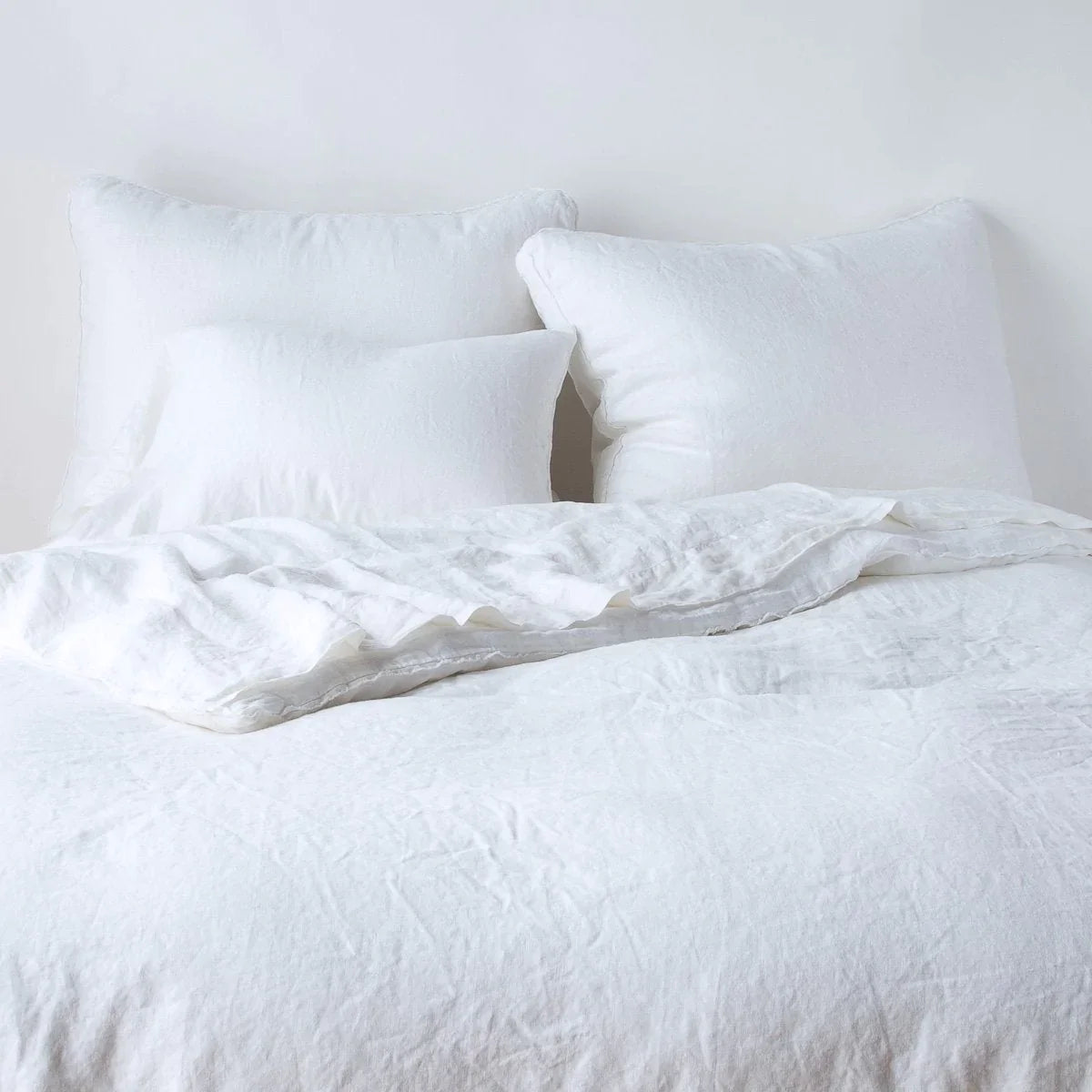 White: Midweight linen shams shown from foot of bed, leaning against a plain background with monochromatic bedding. 