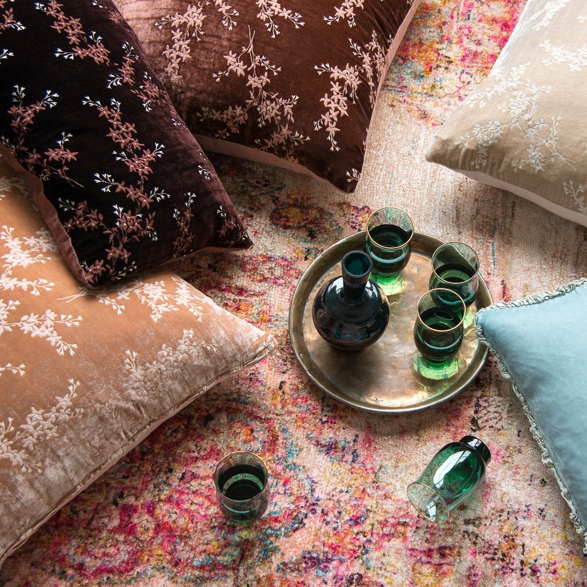 : Lynette throw pillows in pink and brown tones scattered on a bright patterned rug, shown with light blue silk velvet, metal tray, and green glassware - overhead view.