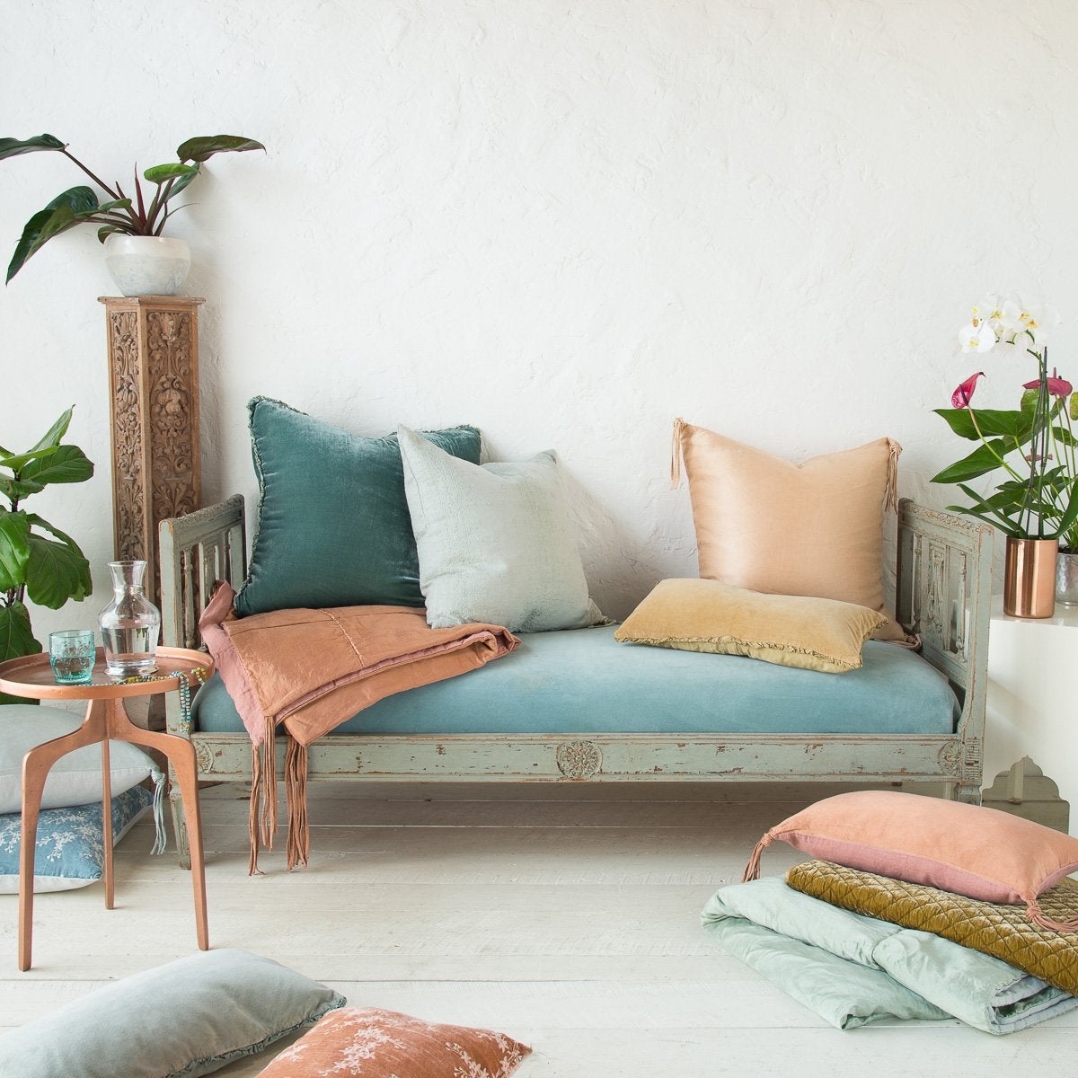 : Ines 24 by 24 pillow on an soft blue antique day bed, layered with accessory pieces in pink, blue, and green tones in linen and charmeuse. Styled with copper accents and greenery. 