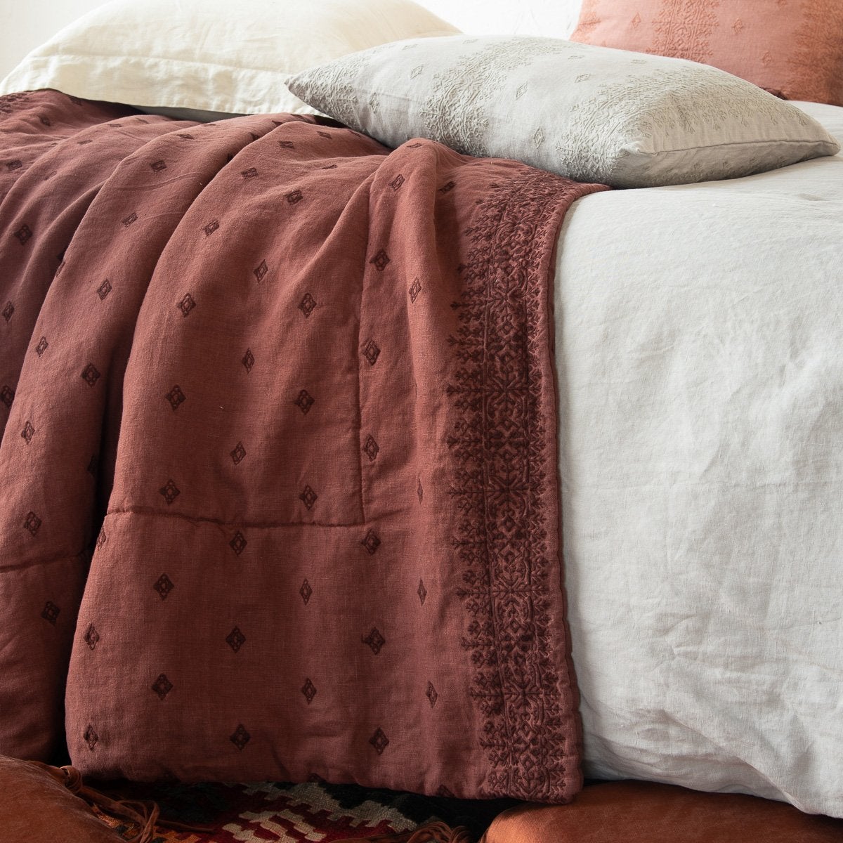 : Ines throw blanket in mahogany rumpled and layered with blue, pink, and cream linen. Close-up side view features cotton embroidery detail.