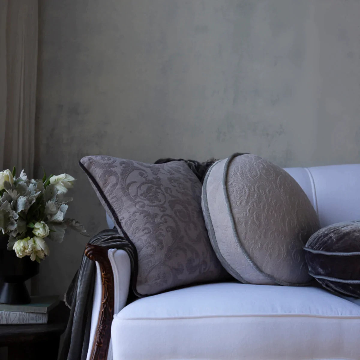 : Square and round pillows on white couch with flowers on side table against a neutral grey wall. 