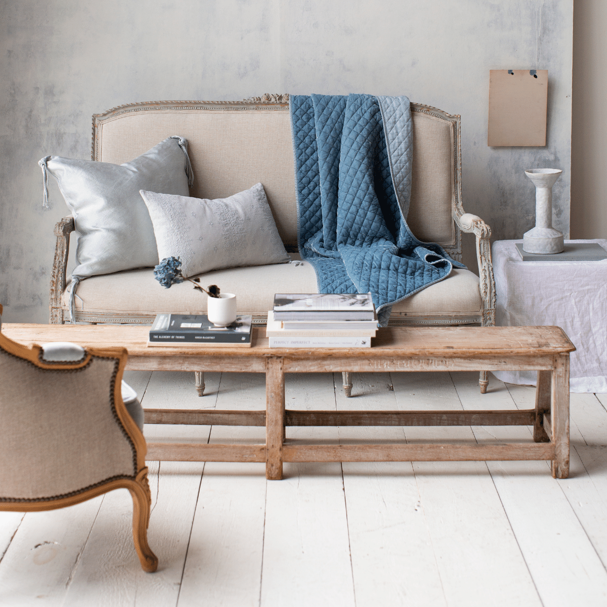[allvariants]: Silk velvet quilted throw blanket in cloud, draped over a light neutral antique couch with light blue throw pillows in silk charmeuse and embroidered linen. Shown with white wood floors, textured light wall, and light wood tones.