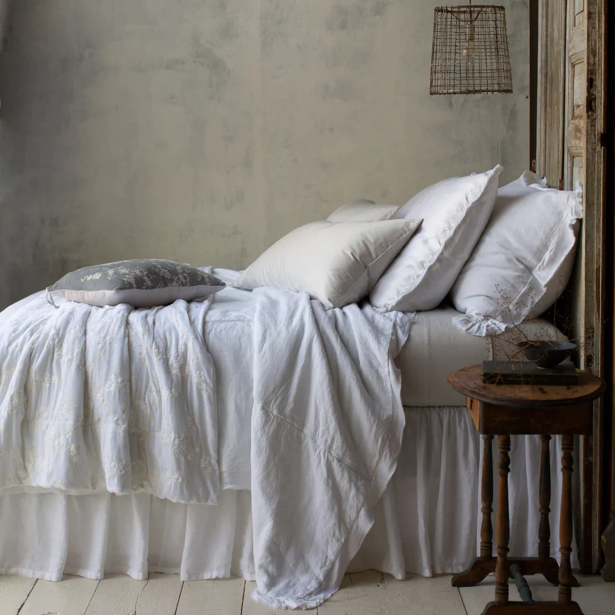 [allvariants]: Sterling Bria sheets contrast against a mostly white bed, shown from the side. The lighting is moody and colors are muted, emphasizing the variety of cotton, linen, and silk textures.