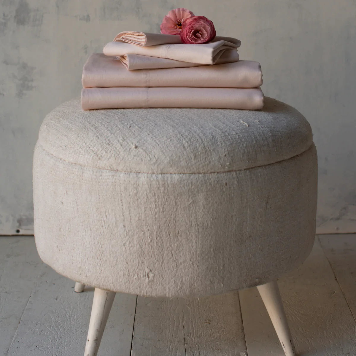 : Soft pink Bria sheets and pillowcases, neatly folded and stacked on a light colored ottoman. Shown from the side, two dark pink flowers rest on top.
