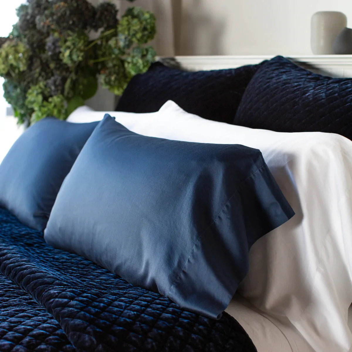 [allvariants]: Bria pillowcases in white and midnight shown from three quarter close up angle. The cotton sateen shines against the deep blue silk velvet quilted shams and coverlet.