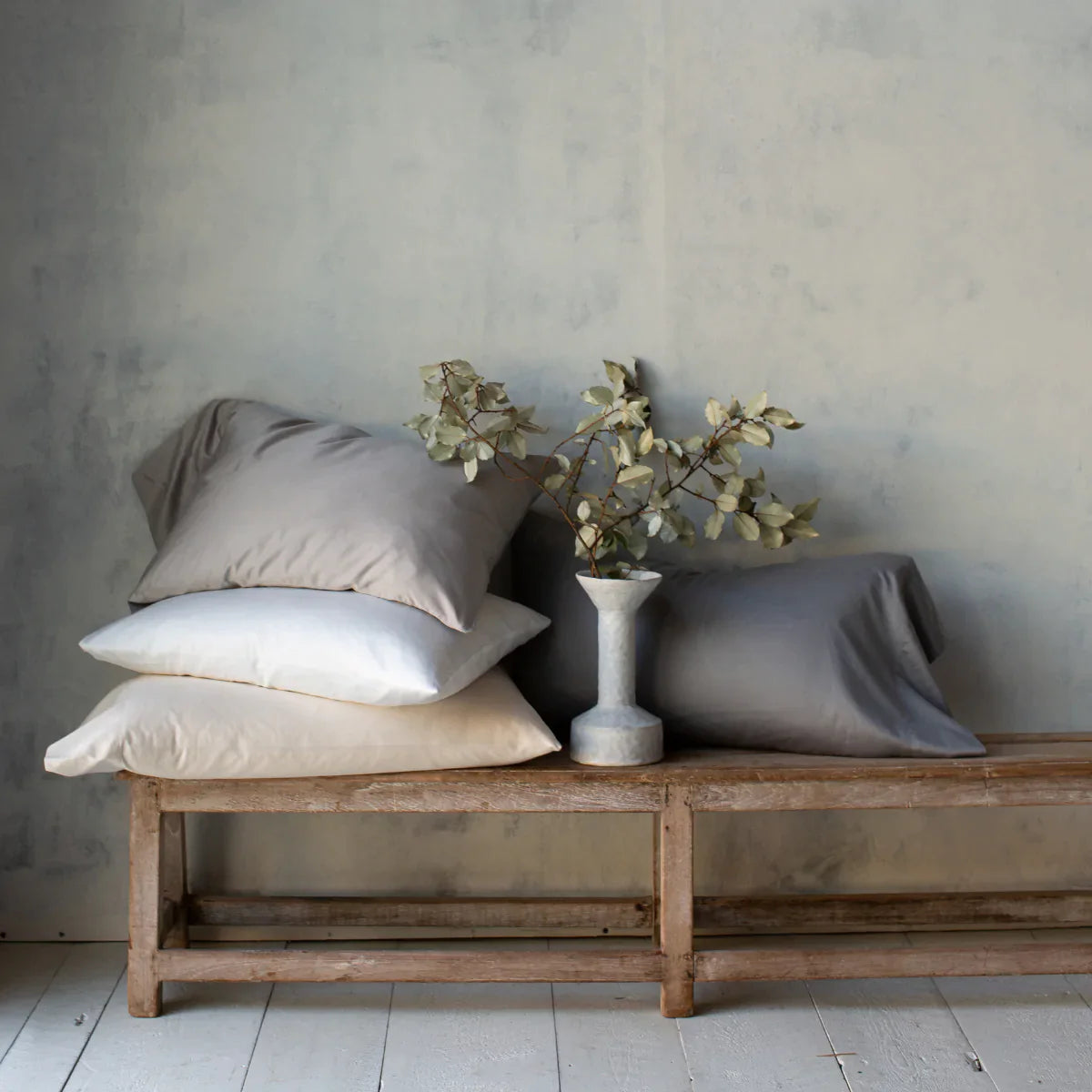 : Bria sleeping pillows in various white and grey tones, stacked on a wooden bench against a textured neutral wall. A ceramic vase with greenery sits between the pillows.
