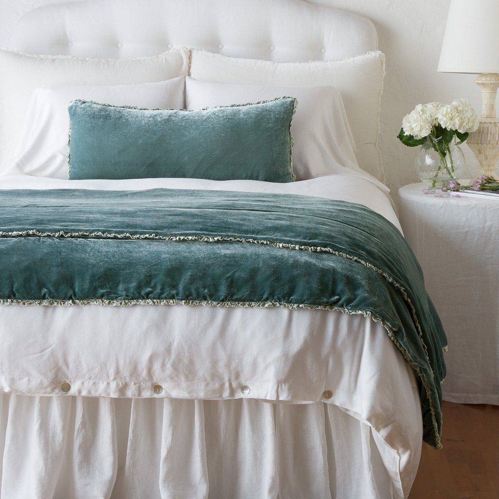 Eucalyptus: Silk velvet lumbar pillow with petite ruffle and throw blanket shown on an all white, neatly made bed - foot of bed angle.