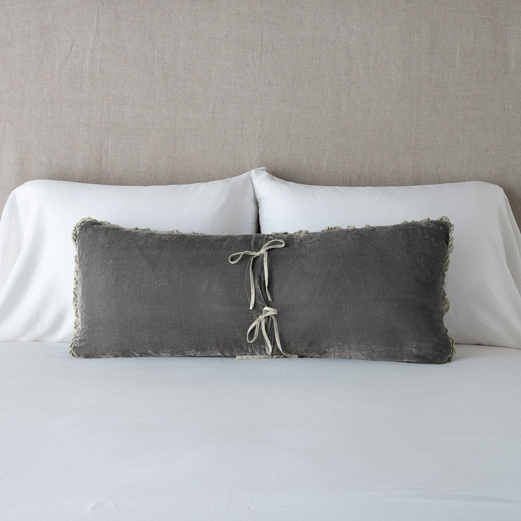 : Carmen lumbar pillow in fog, leaning upright and backwards against neutral bedding and headboard, showcasing the silk charmeuse tie closures.