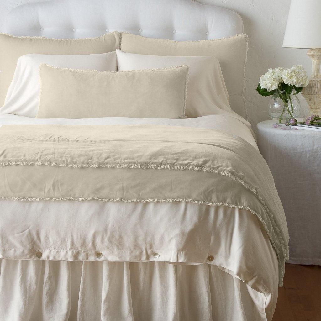 Parchment: Silk velvet lumbar pillow with petite ruffle and throw blanket shown on an all white, neatly made bed - foot of bed angle.