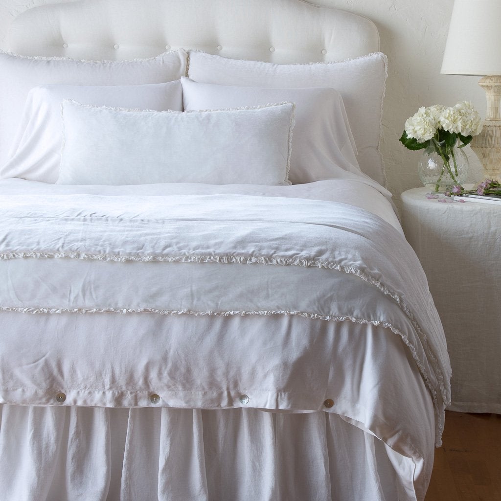 White: Silk velvet lumbar pillow with petite ruffle and throw blanket shown on an all white, neatly made bed - foot of bed angle.