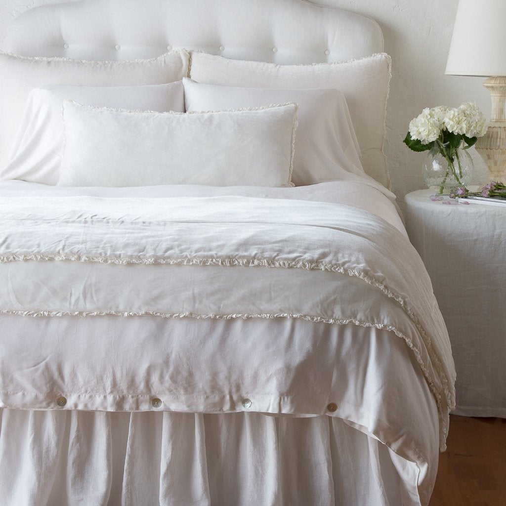 Winter White: Silk velvet lumbar pillow with petite ruffle and throw blanket shown on an all white, neatly made bed - foot of bed angle.