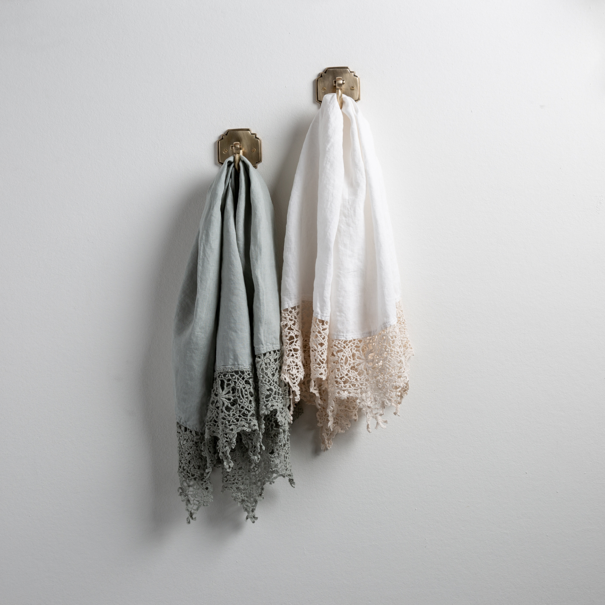 [allvariants]: Two Frida guest towels, draped over decorative hooks against a white wall - eucalyptus and winter white
