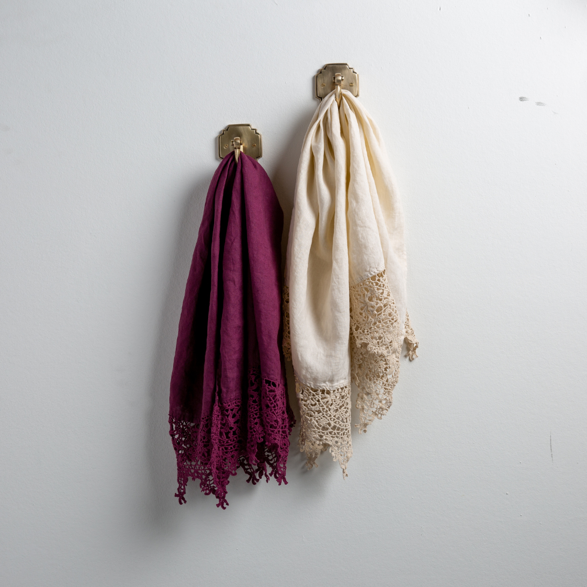 : Two Frida guest towels, draped over decorative hooks against a white wall - fig and parchment.