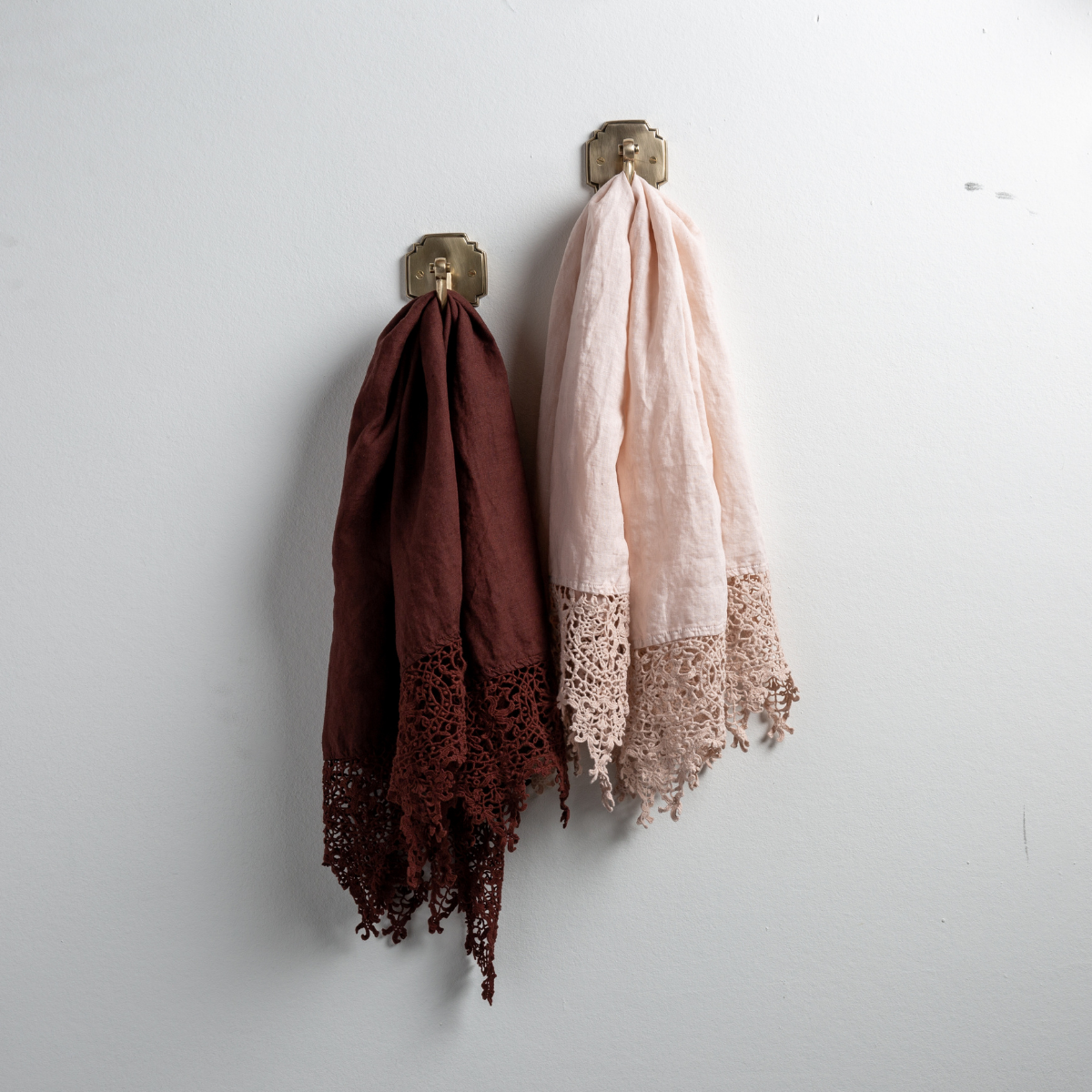 : Two Frida guest towels, draped over decorative hooks against a white wall - mahogany and pearl.