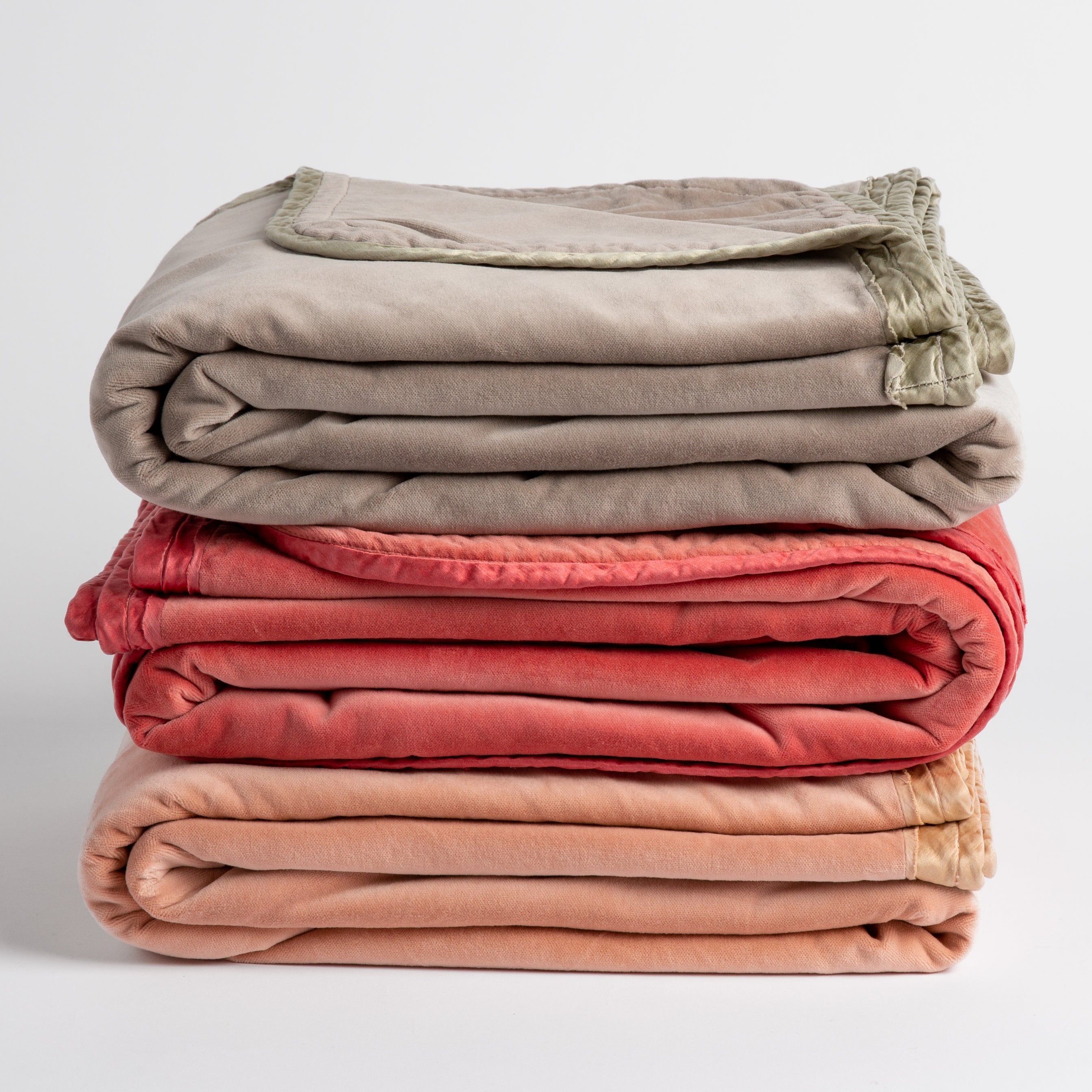 [allvariants]: three cotton velvet throw blankets in fog, poppy and rouge stacked  with charmeuse trim visible. Shot straight on against a plain white background. 