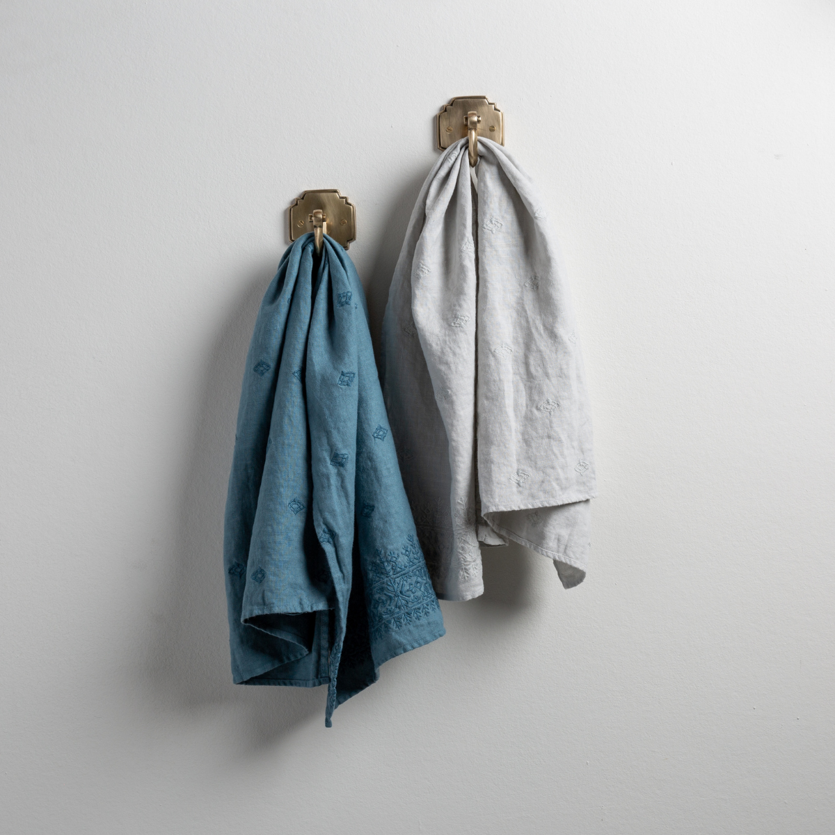 : Two Ines guest towels, draped over decorative hooks against a white wall - cenote and cloud.