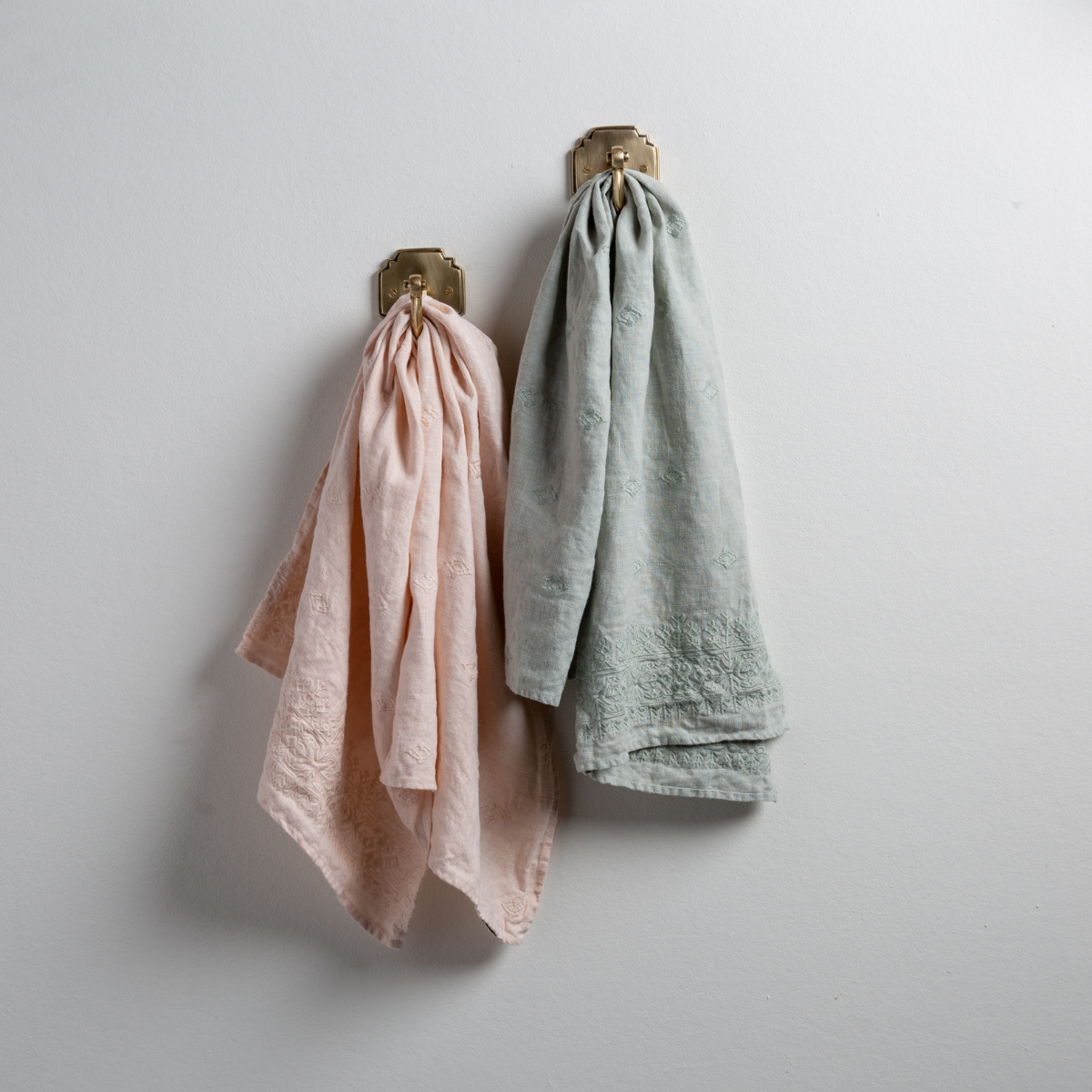 : Two Ines guest towels, draped over decorative hooks against a white wall - pearl and eucalyptus.