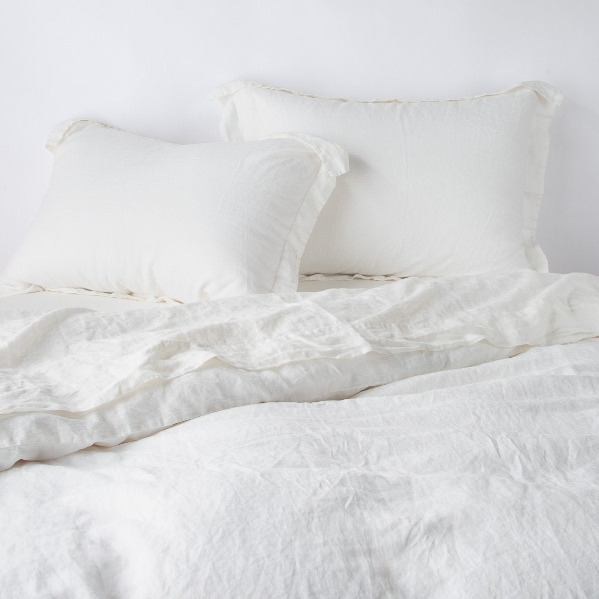 : Linen shams in winter white, leaning upright against a plain wall with matching sheeting and duvet - head-on angle.