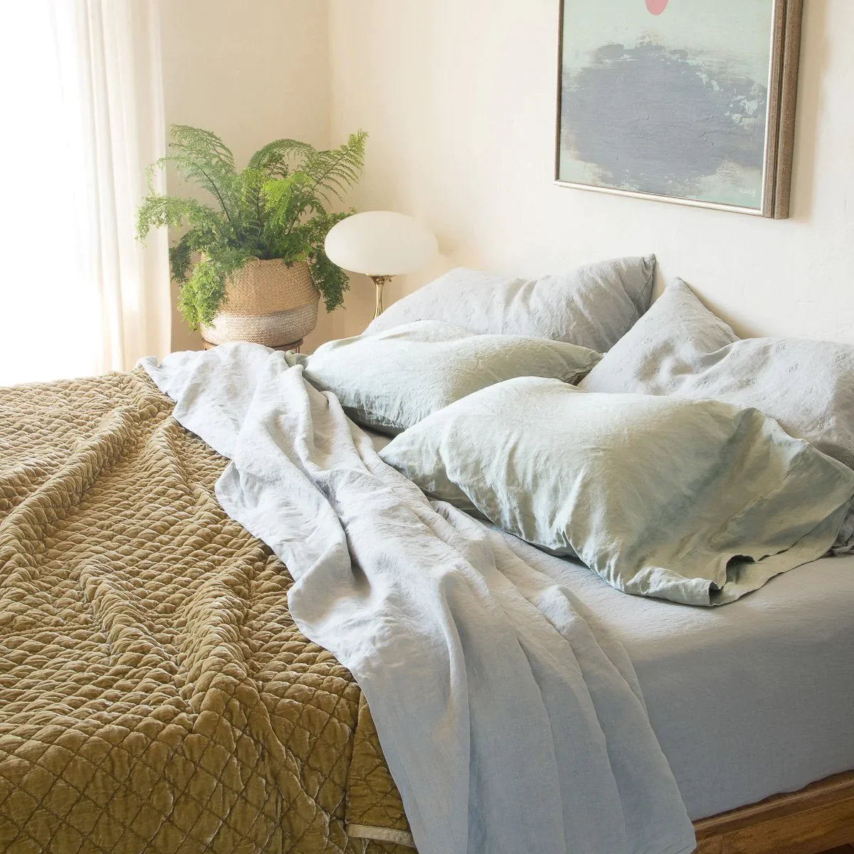[allvariants]: Linen sheeting in cloud with on-tone silk charmeuse sleeping pillows and rich gold toned silk velvet quilted coverlet, styled simply in natural light with a green fern and soft green art - cropped three-quarter angle.