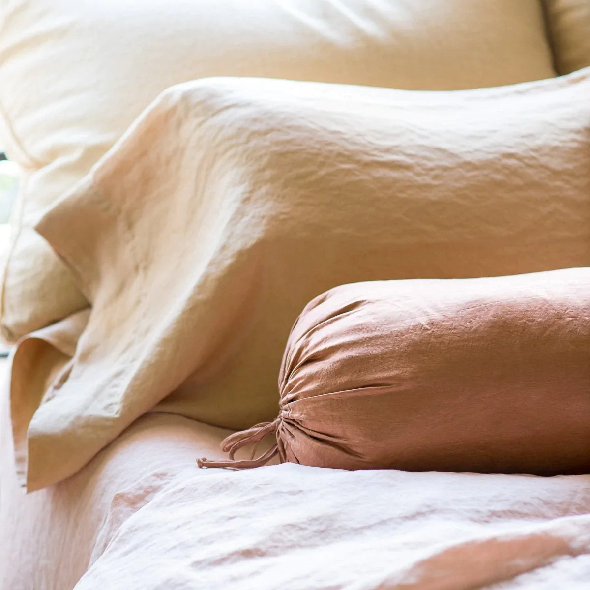 : Paloma bolster and sleeping pillow on linen bedding, in soft pink and gold tones - close-up three-quarter angle.