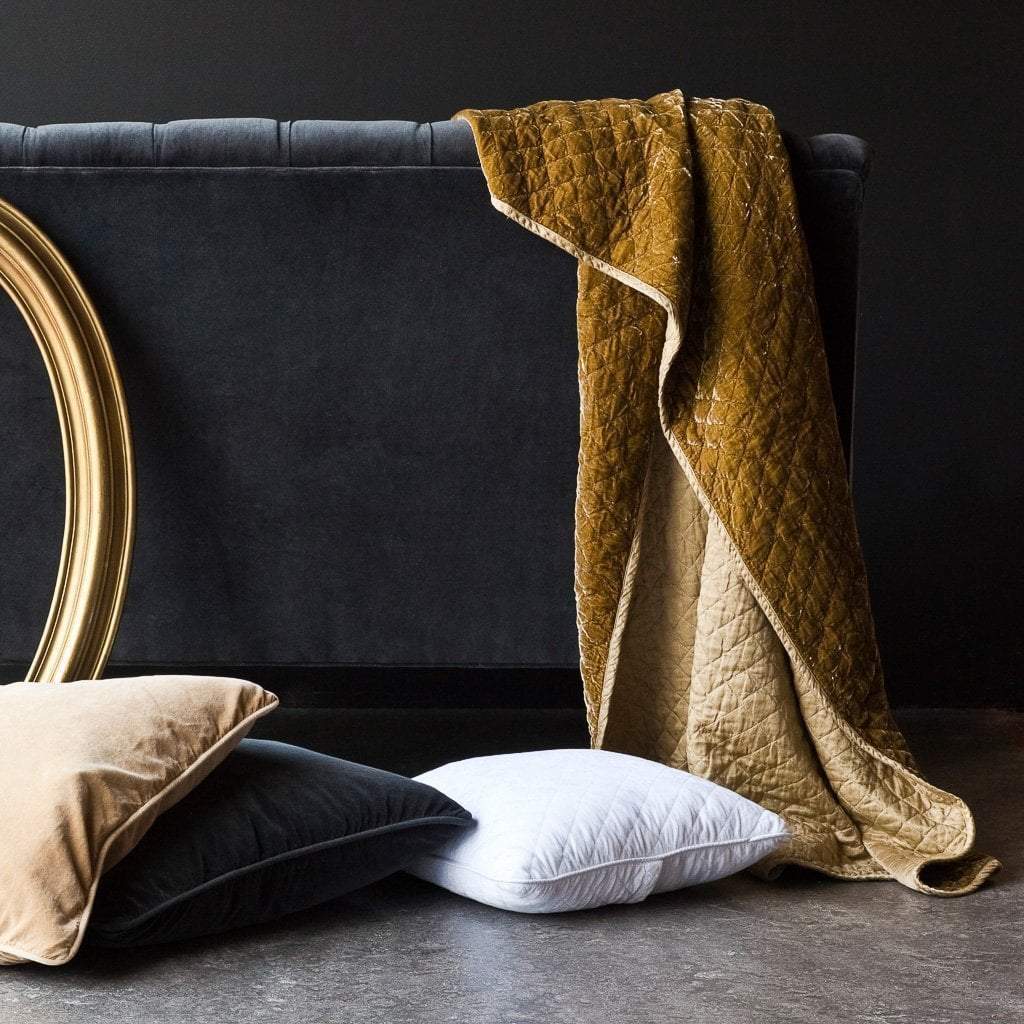 : Silk velvet throw blanket in honeycomb, draped over the back on a dark grey couch. A gold oval frame is partially visible, complementing the rich gold of the blanket.